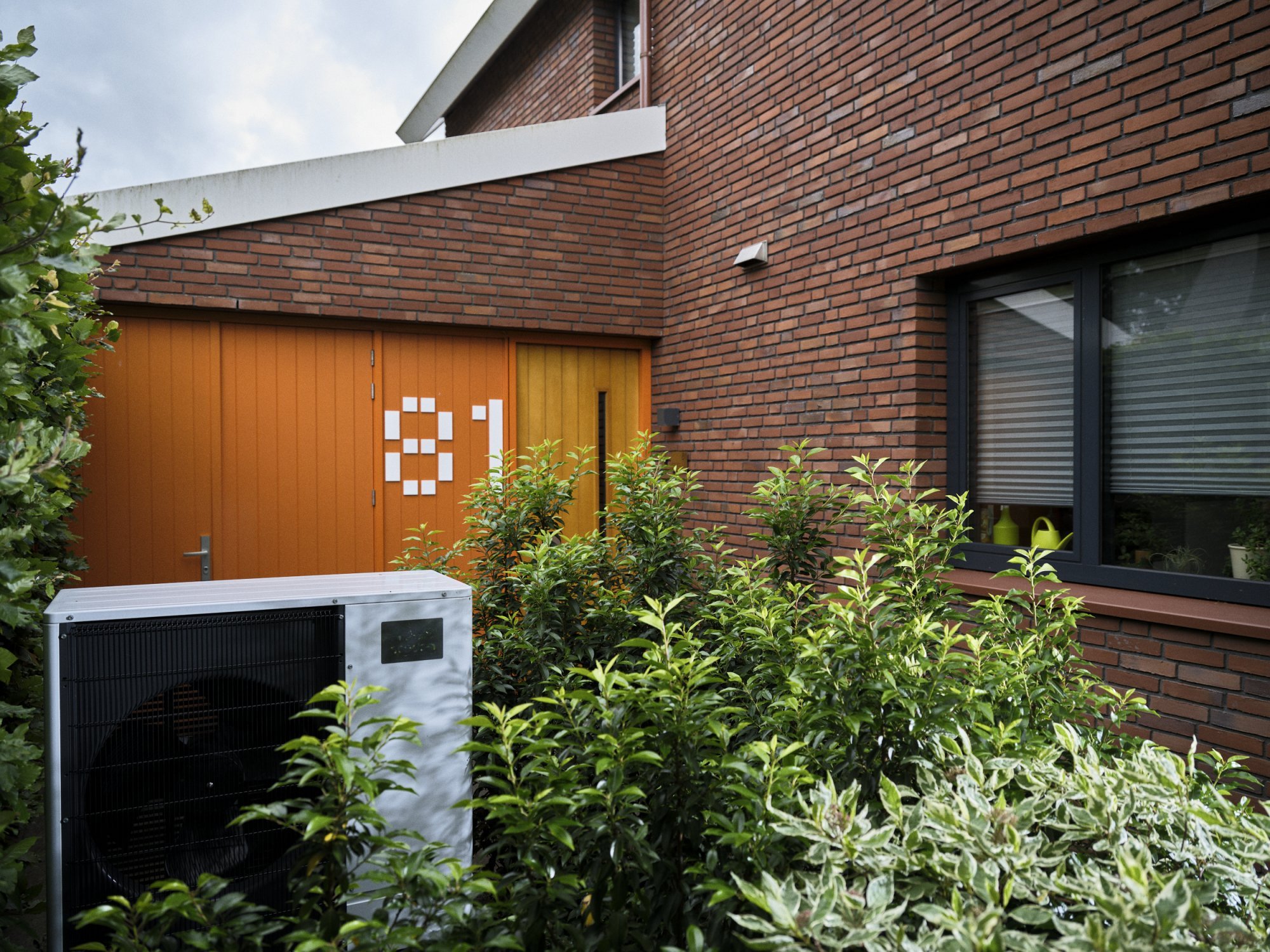 Buitenunit van een warmtepomp naast een huis tussen beplanting
