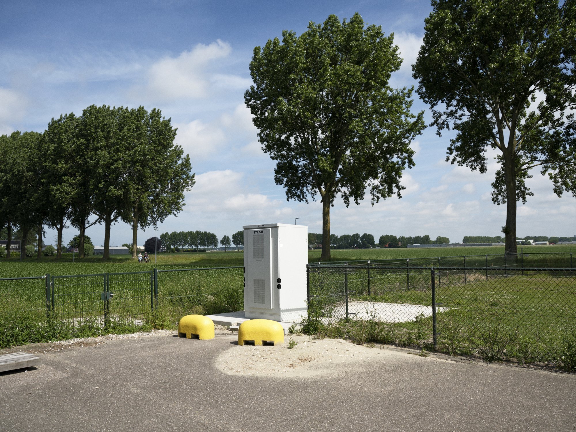 Energiekast langs een landweg, omringd door weiland en bomen.